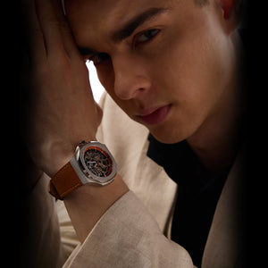 Man wearing a watch with a brown leather strap and a detailed face, looking contemplative.